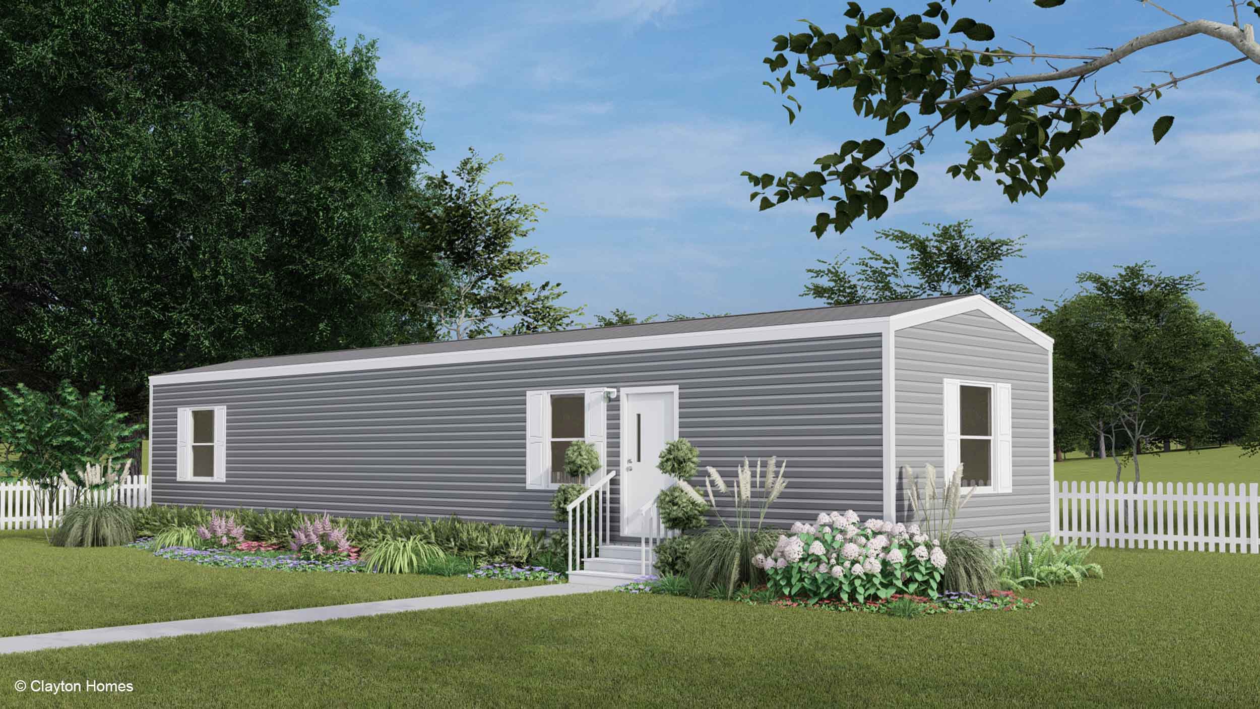 A gray manufactured home with white trim, front steps, garden, and trees sits in a fenced grassy yard.