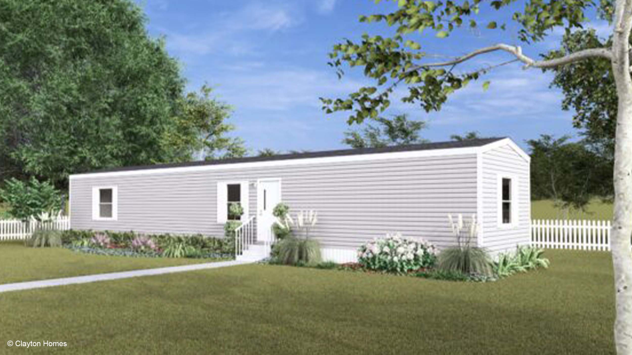 A white single-wide manufactured home with a garden, lawn, and white picket fence sits under a blue sky.