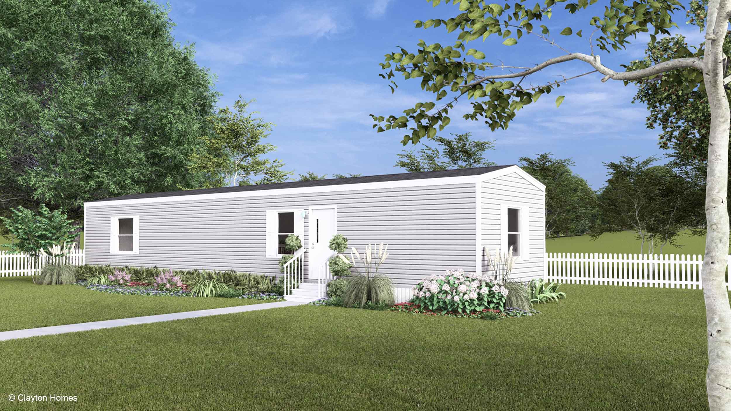 A white manufactured home with steps, a garden, and a white picket fence sits on a grassy yard with trees.